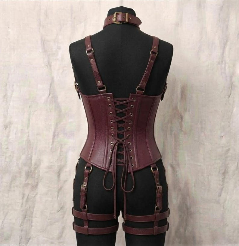 May include: A dark burgundy corset featuring a high neck collar with a buckle, adjustable shoulder straps, and a lace-up back closure. The garment includes attached garter straps with metal buckles that wrap around the upper thighs.