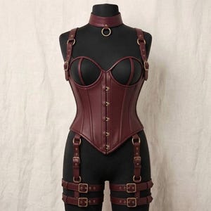 May include: A dark burgundy faux leather corset featuring a sweetheart neckline, front metal hook closures, and an integrated choker with a central ring. The garment includes adjustable shoulder straps and attached thigh garter straps with buckles.