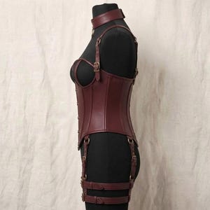 May include: A dark burgundy leather corset featuring a high neck collar, adjustable shoulder straps, and integrated garter straps with metal buckles. The garment is structured with vertical seams and is displayed on a black mannequin torso.