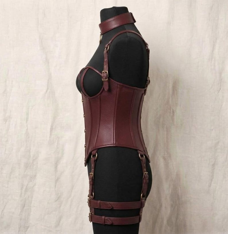 May include: A dark burgundy leather corset featuring a high neck collar, adjustable shoulder straps, and integrated garter straps with metal buckles. The garment is structured with vertical seams and is displayed on a black mannequin torso.