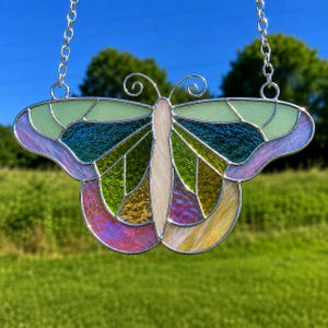 May include: A stained glass butterfly suncatcher with a silver metal frame and chain. The butterfly features a variety of colors, including green, blue, purple, yellow, and white. The wings are divided into geometric shapes.