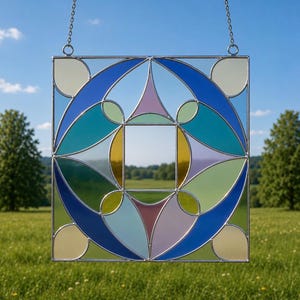 May include: A square stained glass panel with a geometric design, featuring shades of blue, green, yellow, and purple. The panel is suspended by a chain and set against a backdrop of a blue sky and green landscape.