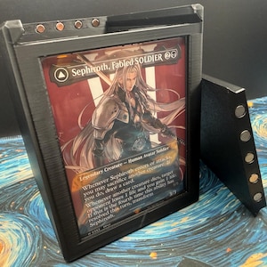 May include: A black card display case with a framed Magic: The Gathering card featuring Sephiroth, Fabled Soldier. The card has a red and white background with an illustration of a character. The case has magnets and a separate stand.