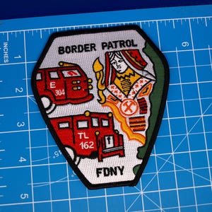 May include: A shield-shaped embroidered patch featuring the text BORDER PATROL and FDNY. The design includes two red fire trucks labeled E 304 and TL 162, and a figure in a red and white robe. It measures approximately 4 inches by 3.5 inches.