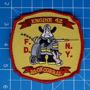 Peut inclure: Écusson jaune en forme de bouclier avec une bordure rouge. Il présente un pompier de dessin animé avec un tuyau, le texte "ENGINE 42", "F.D.N.Y.", et "DA-OK CORRAL" en rouge.