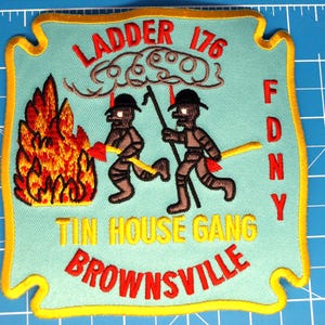 Könnte beinhalten: Gestickter Aufnäher mit hellblauem Hintergrund und gelbem Rand. Der Aufnäher zeigt den Text "LADDER 176", "TIN HOUSE GANG" und "BROWNSVILLE" in Rot und Gelb. Zwei Cartoonfiguren rennen auf ein Feuer zu.