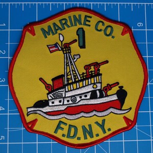Könnte beinhalten: Gestickter Aufnäher mit gelbem Hintergrund und rotem Rand. Der Aufnäher zeigt ein Schlepperdesign mit dem Text "MARINE CO. 1 F.D.N.Y." in Grün. Der Schlepper ist rot, weiß und schwarz, mit einer kleinen amerikanischen Flagge.