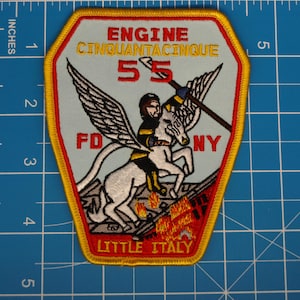 May include: Embroidered patch with a light blue background, featuring a firefighter riding a winged horse. The patch has red, yellow, and black accents, with text that reads "ENGINE CINQUANTACINQUE 55 FD NY LITTLE ITALY". The patch is approximately 10 cm tall.