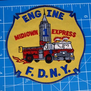 May include: Embroidered patch with a red and white fire engine, the text "ENGINE 1 MIDTOWN EXPRESS F.D.N.Y.", and an illustration of the Empire State Building. The patch has a yellow background with blue text and border.