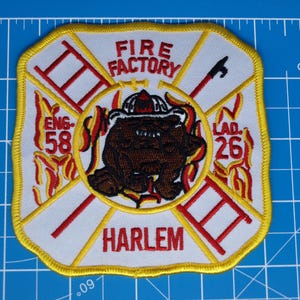 May include: Embroidered patch with a yellow border, featuring a brown bear wearing a crown, surrounded by red ladders and flames. Text includes "FIRE FACTORY," "ENG 58," "LAD 26," and "HARLEM." The patch measures approximately 3.5 inches by 3.5 inches.