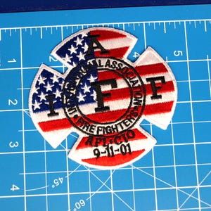 May include: Embroidered patch in the shape of a Maltese cross, featuring the American flag design. The patch includes the text "International Association of Fire Fighters" and "9-11-01".