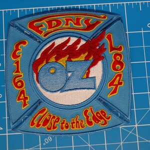 May include: A light blue embroidered patch featuring the text FDNY, E164, L84, and Close to the Edge in red with yellow outlines. The center displays a flame graphic above the letters OZ. The patch is placed on a blue cutting mat with inch markings.