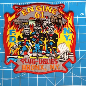 May include: Embroidered patch featuring three firefighters in uniform, with the text "ENGINE 61" and "F.D.N.Y." The patch includes the text "PLUG-UGLIES BRONX, N.Y." and is surrounded by a red and yellow flame design. The patch measures approximately 4 inches by 4 inches.