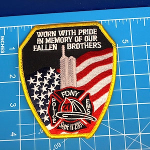May include: A commemorative patch with a yellow border, featuring the text "WORN WITH PRIDE IN MEMORY OF OUR FALLEN BROTHERS." The patch includes an American flag, the Twin Towers, and the FDNY logo. The text "Sept 11 2001" is also present.