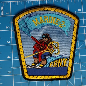 May include: A shield-shaped embroidered patch featuring a firefighter on skis spraying water from a hose. The patch displays the text MARINE-2 in yellow at the top and F.D.N.Y. in yellow at the bottom, with a yellow rope-style border on a blue background.