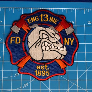 Puede incluir: Parche bordado con un diseño de bulldog, el texto "ENG 13 INE" y "FD NY". El parche tiene forma de cruz de Malta con hachas rojas y el texto "est. 1895". El parche tiene una combinación de colores azul, rojo, naranja, gris y amarillo.