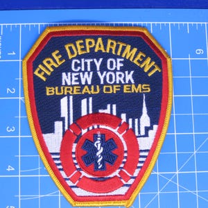 May include: A shield-shaped embroidered patch featuring the text FIRE DEPARTMENT CITY OF NEW YORK BUREAU OF EMS in yellow and white. It displays a white city skyline, a red life preserver, and a blue Star of Life symbol on a dark blue background.