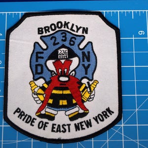 Peut inclure: Un écusson blanc avec une bordure noire représentant un pompier de dessin animé avec une barbe rouge et des bouteilles jaunes. L'écusson porte l'inscription "BROOKLYN 236" et "PRIDE OF EAST NEW YORK" en texte noir. L'écusson mesure environ 10 cm de haut et 10 cm de large.