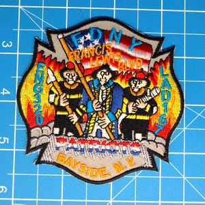 Peut inclure: Écusson brodé sur le thème des pompiers. L'écusson a la forme d'un bouclier avec un motif de flamme. Il comprend le texte "FDNY FRANCIS LEWIS BLVD" et "BAYSIDE N.Y." Trois personnages sont représentés, dont un tenant un drapeau. L'écusson est sur un fond quadrillé bleu.