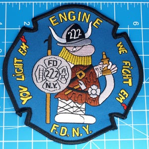Pode incluir: Um patch azul com um bombeiro viking de desenho animado segurando um escudo e um extintor. O patch tem o texto "ENGINE 222" e "F.D.N.Y.", com as frases "YOU LIGHT EM" e "WE FIGHT EM" nas bordas.