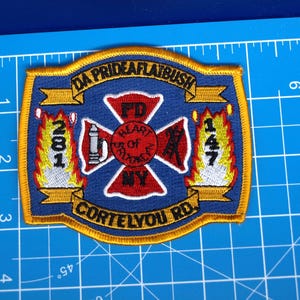 Peut inclure: Écusson brodé avec un fond bleu et une bordure dorée. L'écusson comporte le texte "DA PRIDEAFLATBUSH" et "CORTELYOU RD." ainsi que les chiffres "281" et "147". Une croix rouge et une borne-fontaine blanche sont au centre. L'écusson mesure environ 7.6 cm de large.