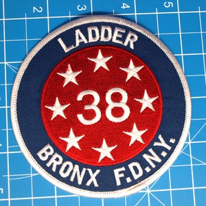 May include: A round patch with the text "LADDER BRONX F.D.N.Y." in white lettering on a navy blue background. The center features a red circle with the number "38" and white stars.
