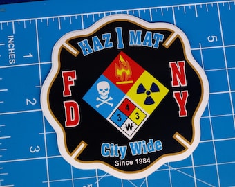 Op de afbeelding: Een zwart, wit, rood, blauw en geel embleem in de vorm van een Maltezer kruis. Het bevat de tekst HAZ MAT, FD NY, City Wide en Since 1984. Een centrale ruit toont een vlam, schedel, stralingssymbool en de gevarencijfers 4, 3, 3, W.