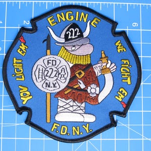 May include: A blue embroidered patch featuring a cartoon character wearing a helmet labeled 222, holding a shield with FD 222 N.Y. and a nozzle. The text reads ENGINE 222, YOU LIGHT EM WE FIGHT EM, and F.D.N.Y. It is placed on a blue mat with a 5-inch scale.