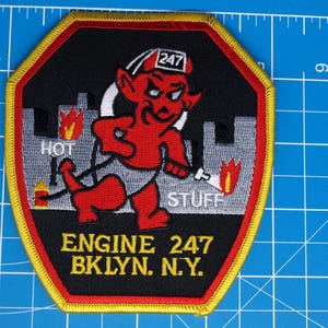 May include: A red, black, and yellow embroidered patch featuring a cartoon devil with the text "HOT STUFF" and "ENGINE 247 BKLYN. N.Y." The patch is approximately 3 inches wide and 4 inches tall.