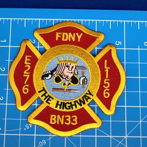 Peut inclure: Écusson brodé FDNY en forme de croix de Malte. Le patch comporte les textes "FDNY", "E276", "L156", "THE HIGHWAY" et "BN33". L'emblème central représente une figure tenant des outils. Le patch est rouge, or et bleu.