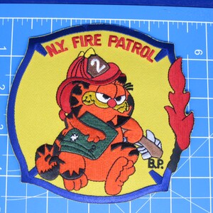 May include: A yellow embroidered patch with a blue border featuring the cartoon character Garfield wearing a red firefighter helmet with the number 2, holding a green book, with the text N.Y. FIRE PATROL and B.P. on a 5-inch grid mat.