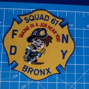 May include: Embroidered patch in a shield shape with a yellow background. The patch features a cartoon firefighter and the text "SQUAD 67", "FDNY", and "BRONX". The text "TAKING IN A JOB NEAR YOU" is also included.