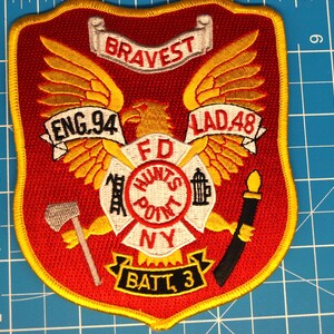 Pode incluir: Emblema bordado vermelho e dourado com o texto "BRAVEST" no topo. O emblema apresenta uma águia com as asas abertas, os textos "ENG.94" e "LAD.48", e as palavras "FD HUNTS POINT NY". O emblema mede aproximadamente 10 cm de altura.