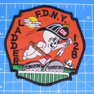 May include: An embroidered F.D.N.Y. Ladder 128 patch featuring a cartoon ghost wearing a firefighter helmet, holding an axe, and flying over tombstones labeled Tombstone Territory and R.I.P. The patch is approximately 4 inches tall.