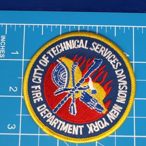 Peut inclure: Un écusson circulaire avec une bordure jaune et un fond bleu. Le texte "CITY OF TECHNICAL SERVICES DIVISION NEW YORK FIRE DEPARTMENT" est en blanc. Le centre présente un cercle rouge avec des outils de pompiers jaunes et bleus.