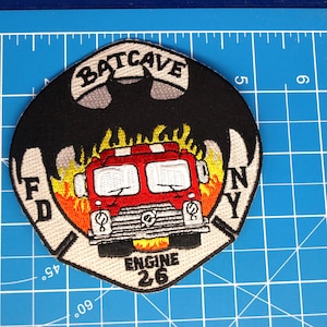 Peut inclure: Écusson brodé représentant un camion de pompiers rouge avec le texte "ENGINE 26" et "BATCAVE" au-dessus. L'écusson présente une silhouette de chauve-souris noire et les lettres "FD NY". Le motif comprend des flammes et est sur une grille bleue.