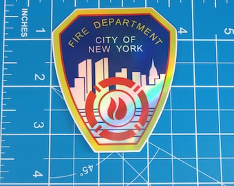 Op de afbeelding: Een holografische sticker met het embleem van de brandweer van New York City. De schildvormige sticker heeft een blauwe achtergrond met de tekst "FIRE DEPARTMENT CITY OF NEW YORK". Het embleem bevat een stadsgezicht en een rood vuursymbool. De sticker is ongeveer 7.6 cm hoog.
