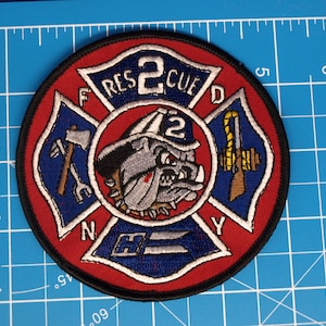 Op de afbeelding: Een ronde geborduurde patch met een rode rand en een centraal ontwerp met een bulldog met een brandweerhelm. De patch bevat de tekst "FIRE RESCUED 2 NY" en een Maltezer kruisontwerp. De patch heeft een diameter van ongeveer 10 cm.