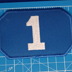 Op de afbeelding: Een rechthoekige blauwe geborduurde patch met afgeschuinde hoeken en een groot wit cijfer één in het midden. De patch ligt op een blauwe snijmat met witte rasterlijnen en inch-markeringen zichtbaar langs de linker- en bovenranden.