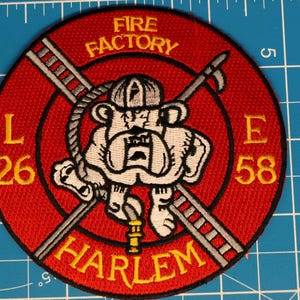 Peut inclure: Un écusson brodé circulaire à fond rouge et bordure noire. Il représente un bouledogue blanc portant un casque de pompier, des échelles croisées et une lance à incendie. Le texte indique FIRE FACTORY, HARLEM, L 26 et E 58 en fil jaune.