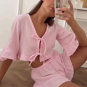 May include: A light pink two-piece set consisting of a cropped top and shorts. The top features a V-neckline, three-quarter length ruffled sleeves, and two front tie closures. The matching shorts have an elastic waistband and a ruffled hem.