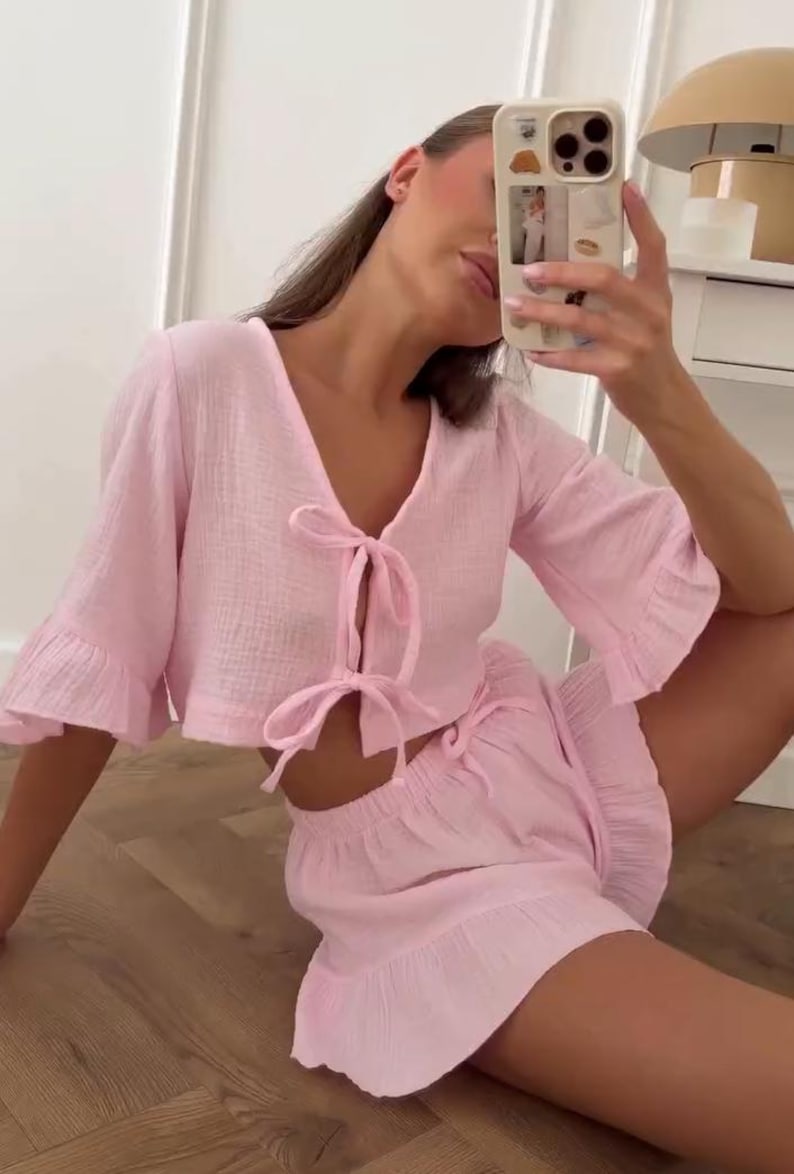May include: A light pink two-piece set consisting of a cropped top and shorts. The top features a V-neckline, three-quarter length ruffled sleeves, and two front tie closures. The matching shorts have an elastic waistband and a ruffled hem.