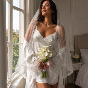 May include: A bride in a white satin slip dress with lace detailing, a matching robe, and a sheer veil. She holds a bouquet of white calla lilies. The scene is set near a window with natural light.