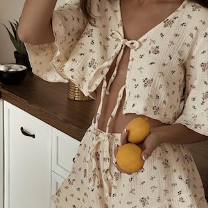 May include: A two-piece set consisting of a cropped top and shorts in a cream-colored fabric with a small floral print. The top features a V-neckline with a front tie closure and short flared sleeves. The shorts have a drawstring waist and ruffled hems.