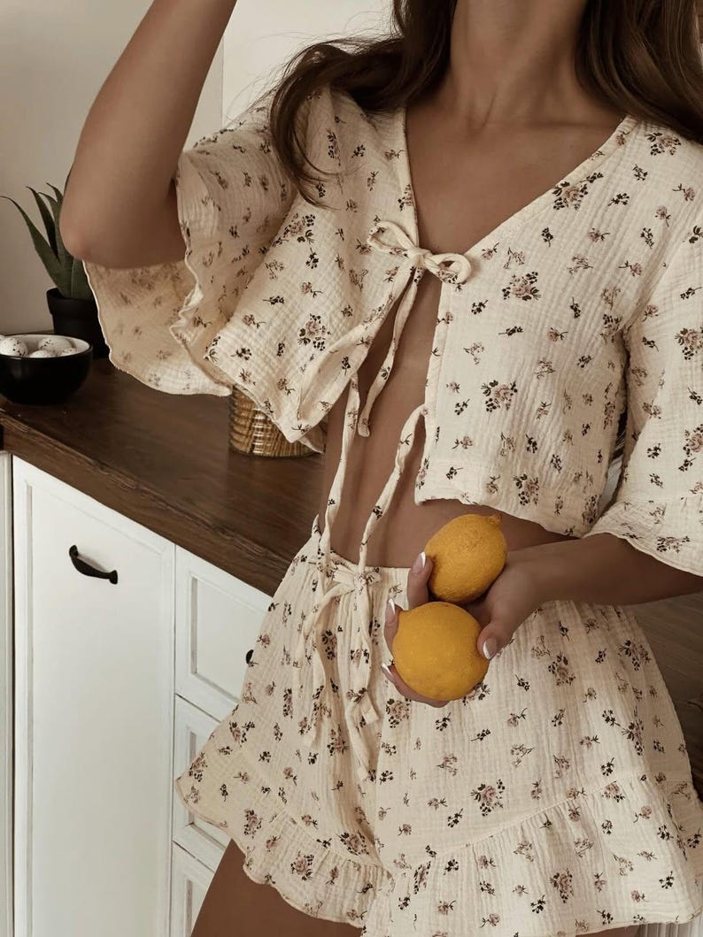 May include: A two-piece set consisting of a cropped top and shorts in a cream-colored fabric with a small floral print. The top features a V-neckline with a front tie closure and short flared sleeves. The shorts have a drawstring waist and ruffled hems.