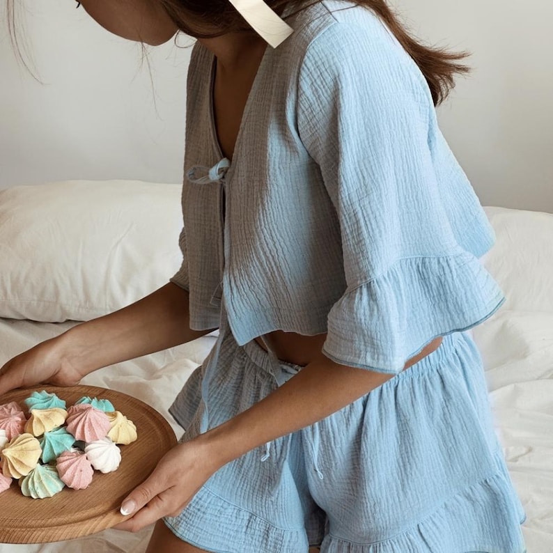 May include: A light blue two-piece set consisting of a cropped top with a V-neckline, front tie closure, and ruffled sleeves, paired with matching ruffled shorts. The fabric has a textured, crinkled appearance throughout the entire garment.