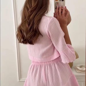 May include: A light pink two-piece set consisting of a long-sleeved, textured crop top and matching high-waisted shorts with a ruffled hem. The fabric has a visible crinkled, gauze-like texture, and the shorts feature an elasticized waistband.