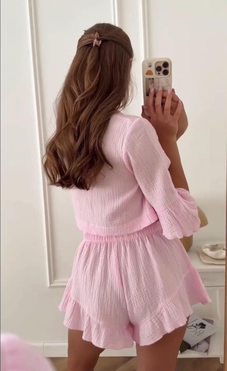 May include: A light pink two-piece set consisting of a long-sleeved, textured crop top and matching high-waisted shorts with a ruffled hem. The fabric has a visible crinkled, gauze-like texture, and the shorts feature an elasticized waistband.