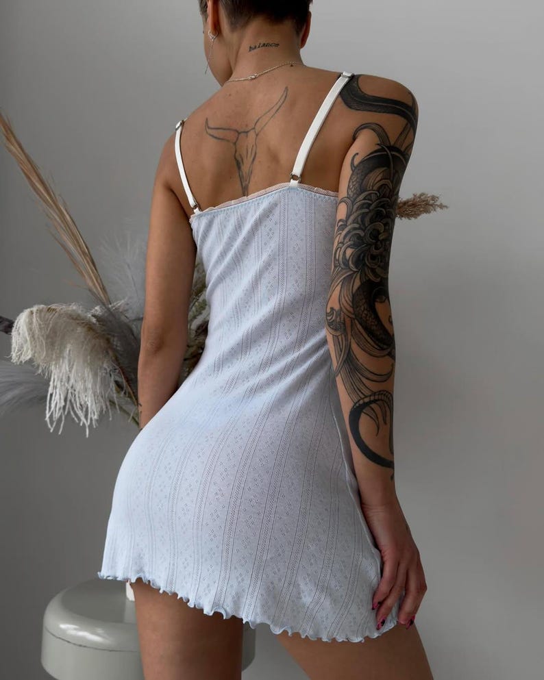 Puede incluir: Un vestido lencero azul claro con tirantes blancos y un dobladillo festoneado. El vestido tiene un sutil estampado floral y una textura acanalada. Las correas son ajustables. El vestido es de largo mini.
