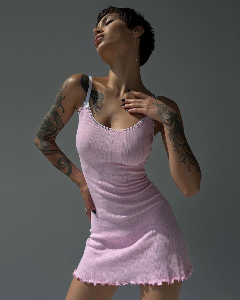 Puede incluir: Vestido lencero rosa claro de punto acanalado con ribete y tirantes blancos. El vestido tiene un dobladillo festoneado y una silueta ajustada. La modelo lleva el vestido. El fondo es gris liso.